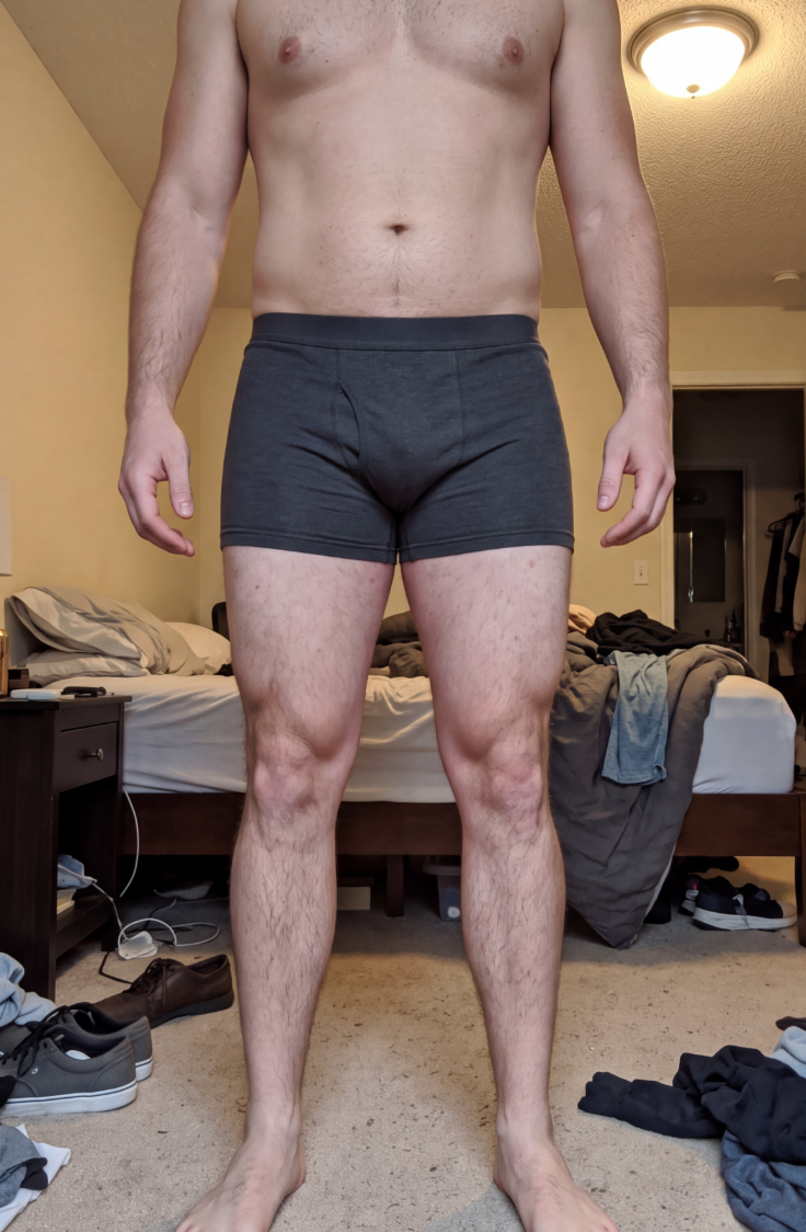 A muscular man stands in his boxer briefs in a messy bedroom.