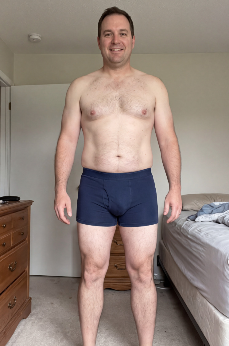 A smiling, shirtless man in blue boxer briefs stands facing forward in a bedroom.