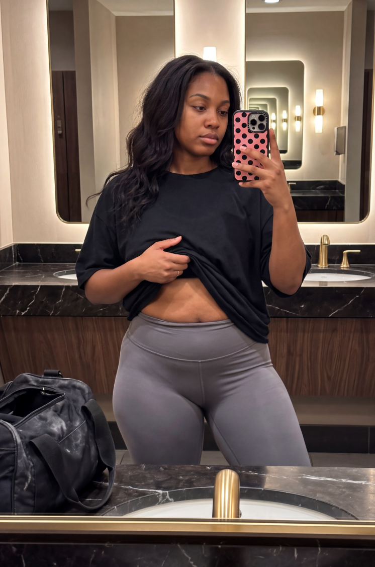 A woman in a black bikini looks at her reflection in a bathroom mirror.
