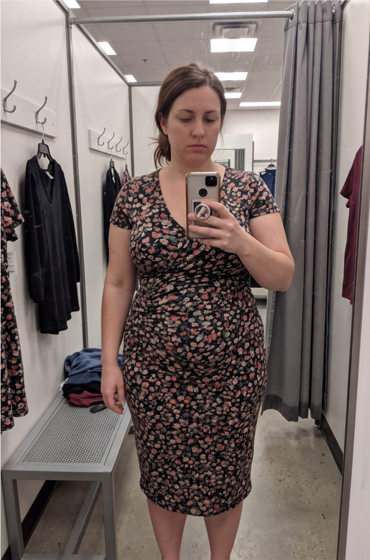 A woman takes a mirror selfie while wearing a short-sleeved, floral-print dress in a fitting room.