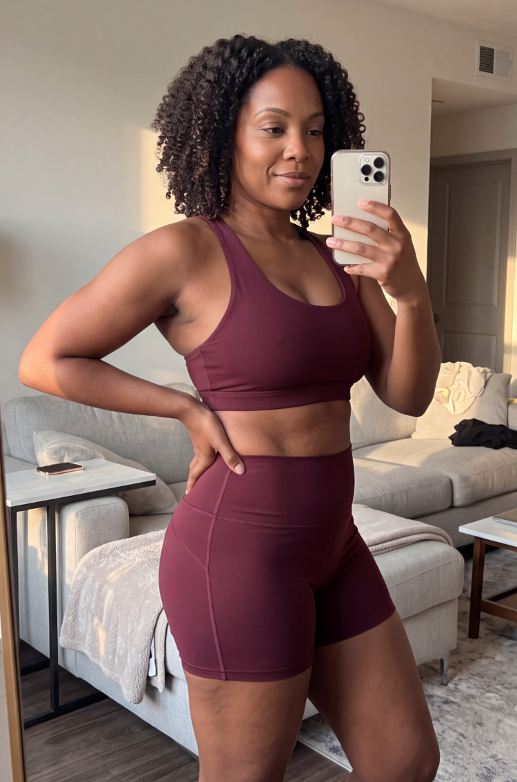 A woman in a matching maroon sports bra and athletic shorts takes a mirror selfie indoors.