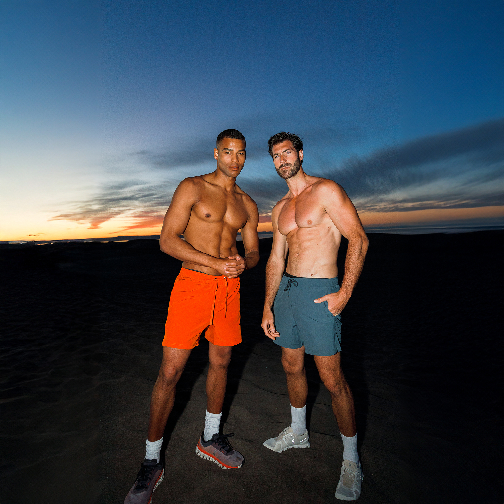 Two men in shorts standing on a beach at sunset.