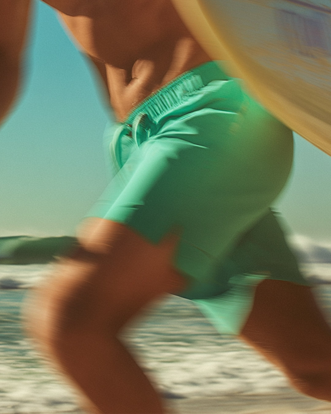 Person in green shorts running on a beach with a surfboard.