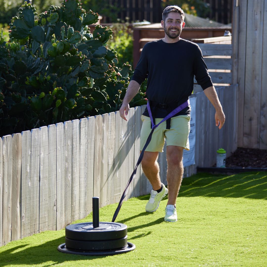 Man pulling weighted sled on green grass near wooden fence.