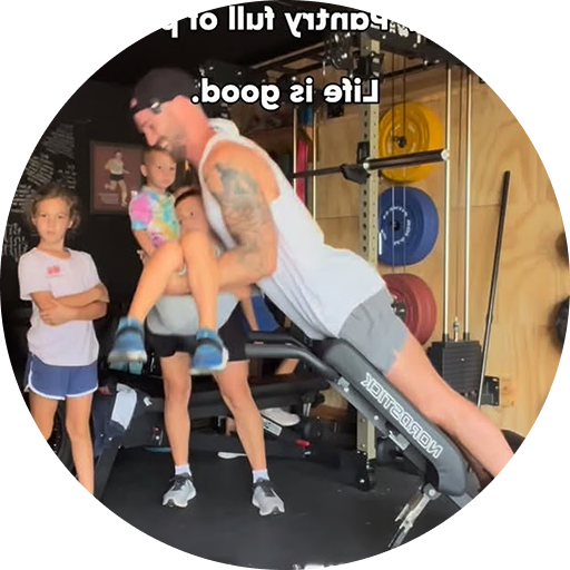 A man in a home gym exercises on a bench while holding a child, as two other children watch.