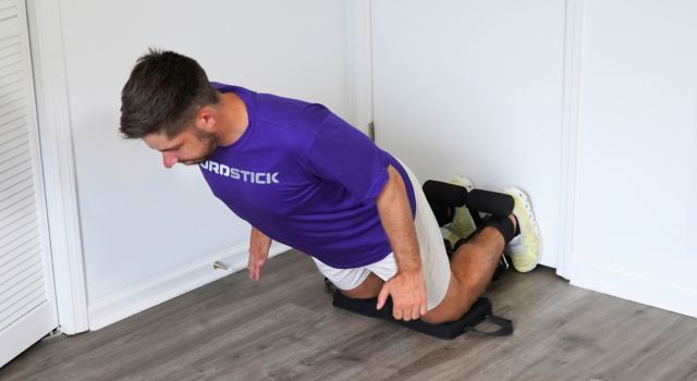 Man in purple shirt performing a kneeling exercise on a wooden floor.