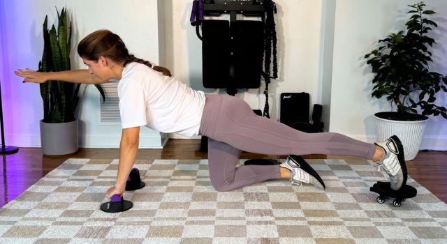 Woman exercising on a mat, extending one arm and opposite leg, with plants in the background.