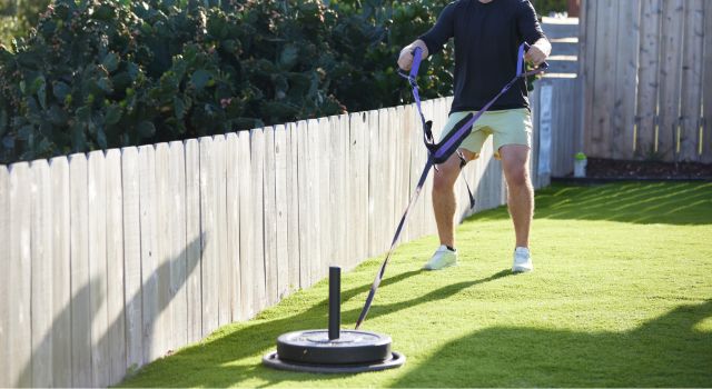 Person pulling a weighted sled on grass in a backyard setting.