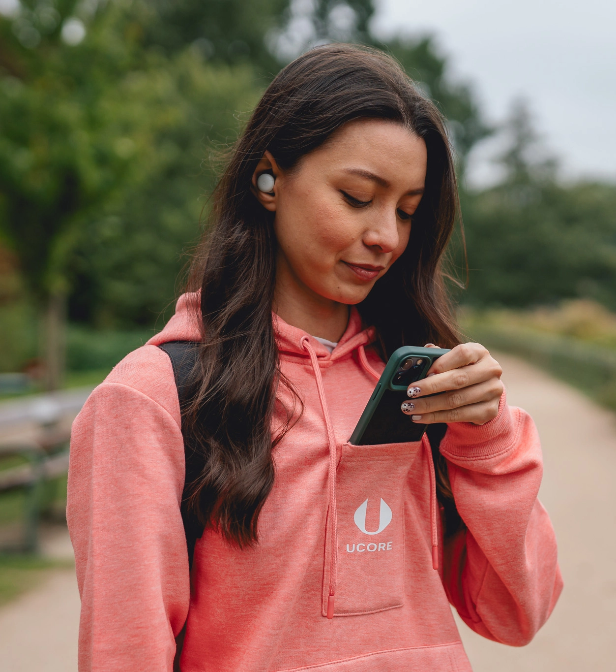 Woman in pink hoodie using smartphone outdoors, wearing earbuds.