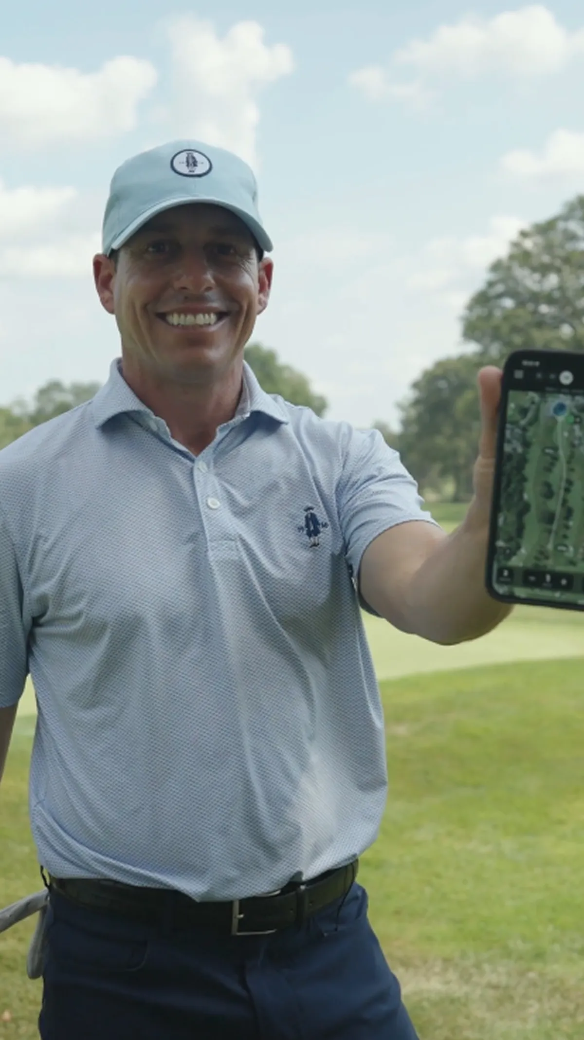 A smiling man on a golf course holds up a phone showing a map of the hole.