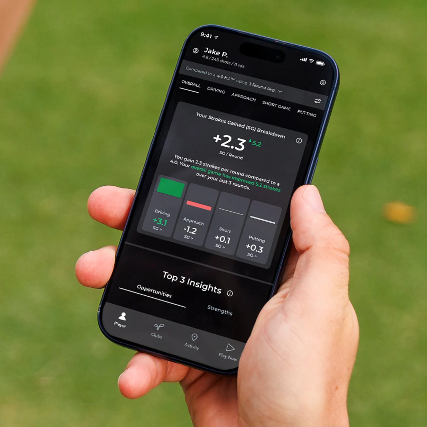 A person holds a smartphone displaying a golf statistics app against a green grass background.