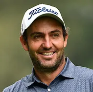 A smiling man with a beard wearing a white Titleist baseball cap and a collared shirt.