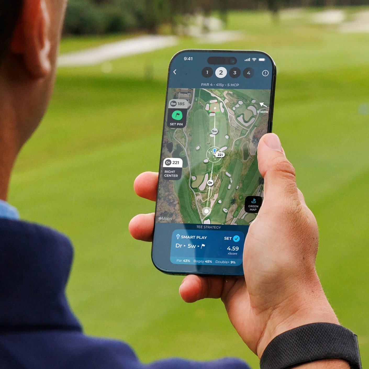 A person holds a smartphone displaying a golf course map on an app, with a green lawn behind.