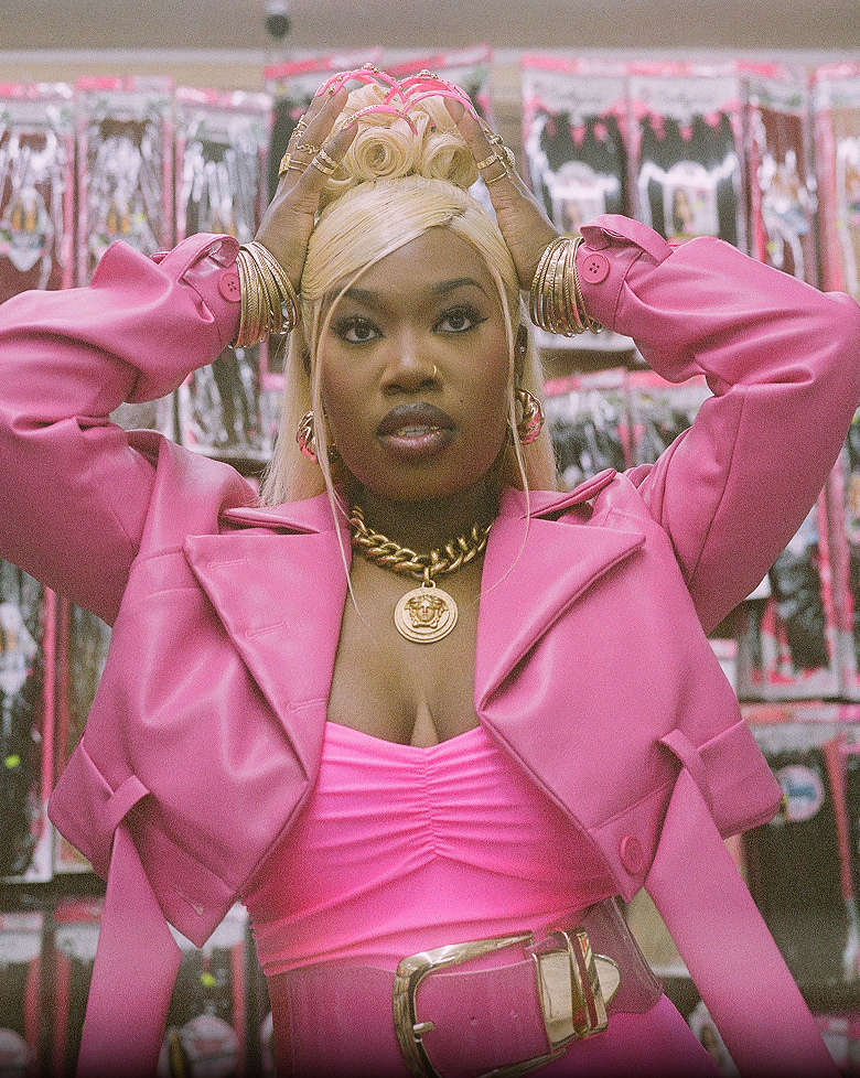 A woman in a pink outfit holds her hands up to her elaborate blonde updo.