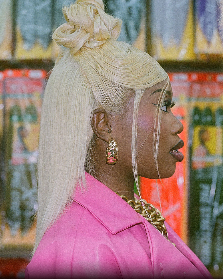 Side profile of a woman with long blonde hair in a half-up bun, wearing a pink jacket.
