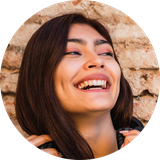 A circular photo of a young woman with long brown hair, smiling and looking up against a brick wall.