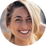 A circular, close-up photo of a smiling woman with blonde hair, with her hand in her hair.