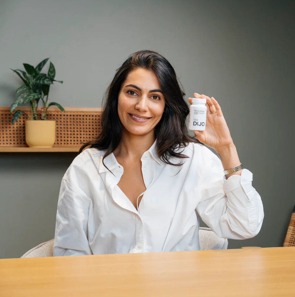 A smiling woman in a white shirt sits at a table holding a small white bottle labeled DIJO.