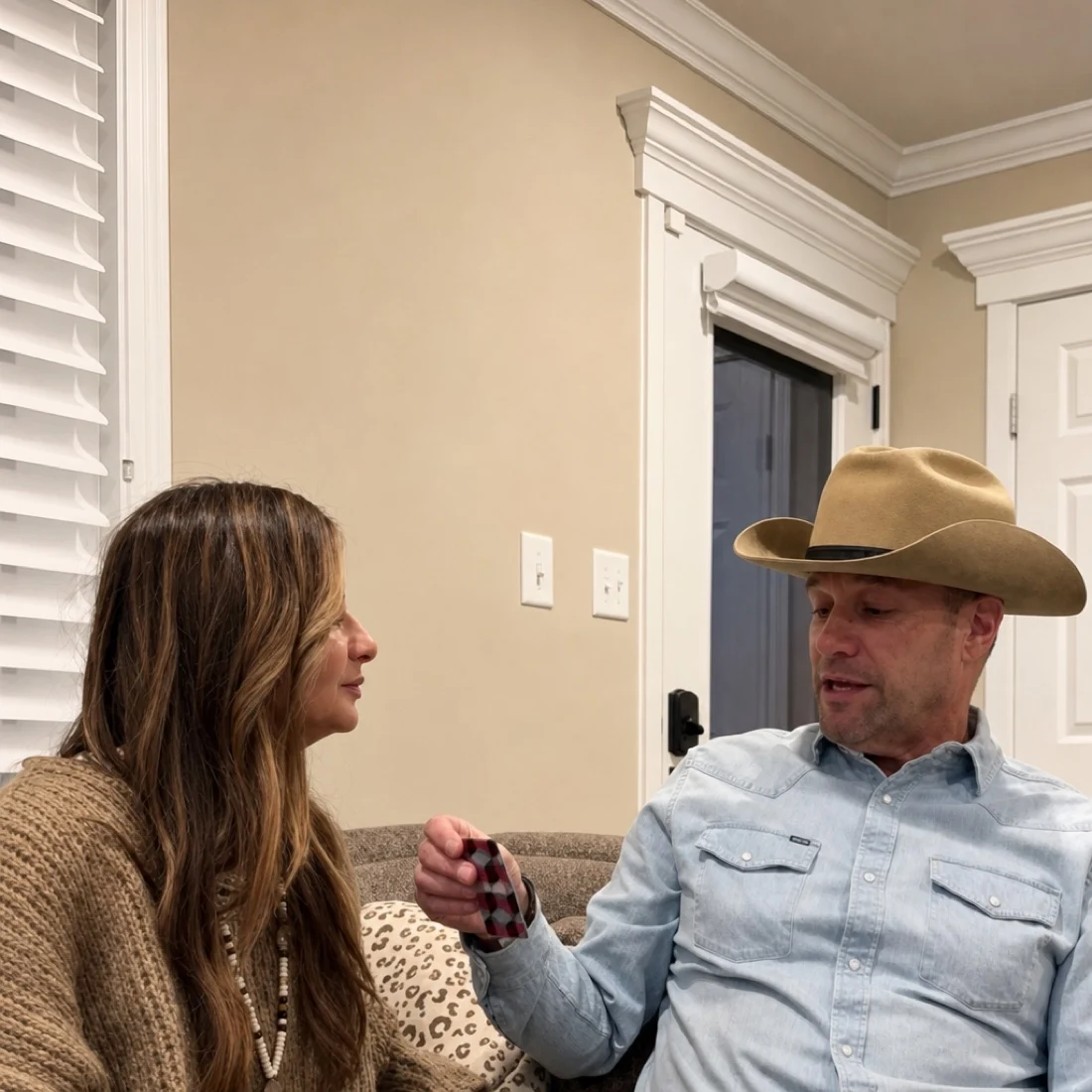 A man in a cowboy hat and a woman in a sweater sit on a couch talking.