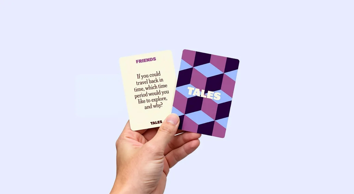 A hand holding two cards from the game 'Tales' against a plain light purple background.