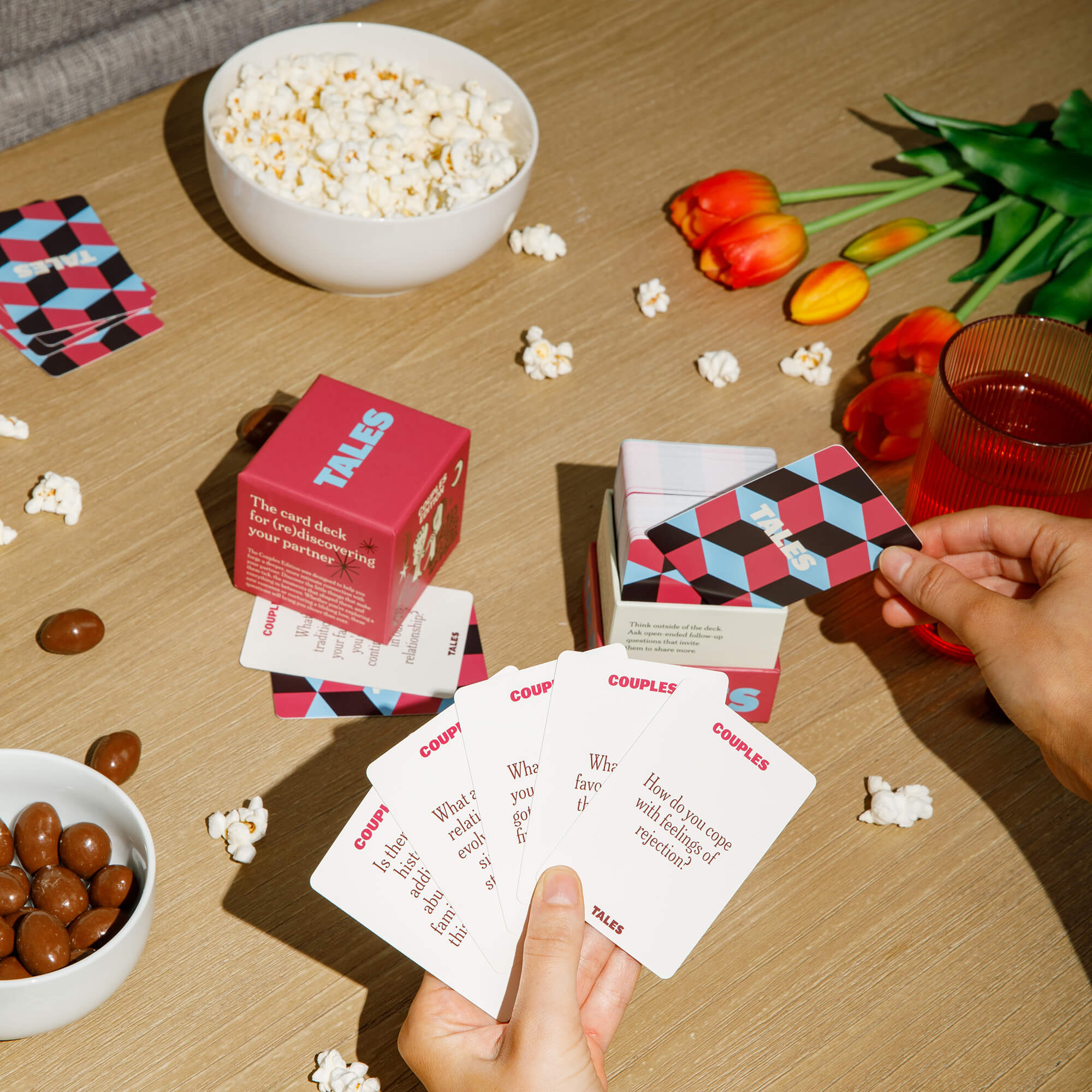 Hands holding cards, popcorn, chocolates, tulips, and a red drink on a table.