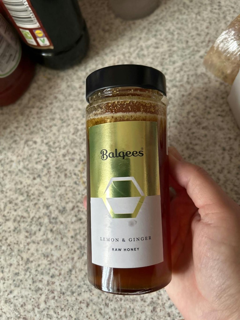 A person's hand holding a jar of Balqees Lemon & Ginger Raw Honey against a kitchen counter.