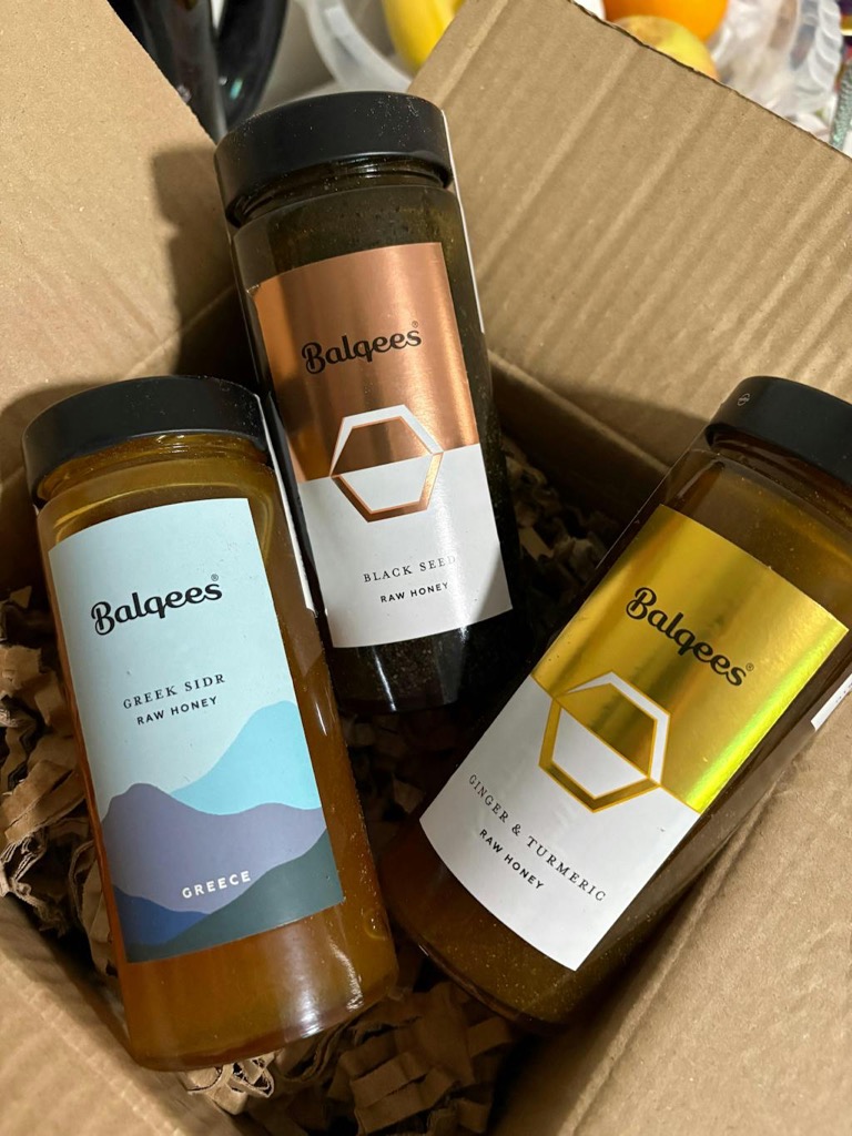Three jars of Balqees raw honey, in different flavors, sitting inside a cardboard box with packing material.