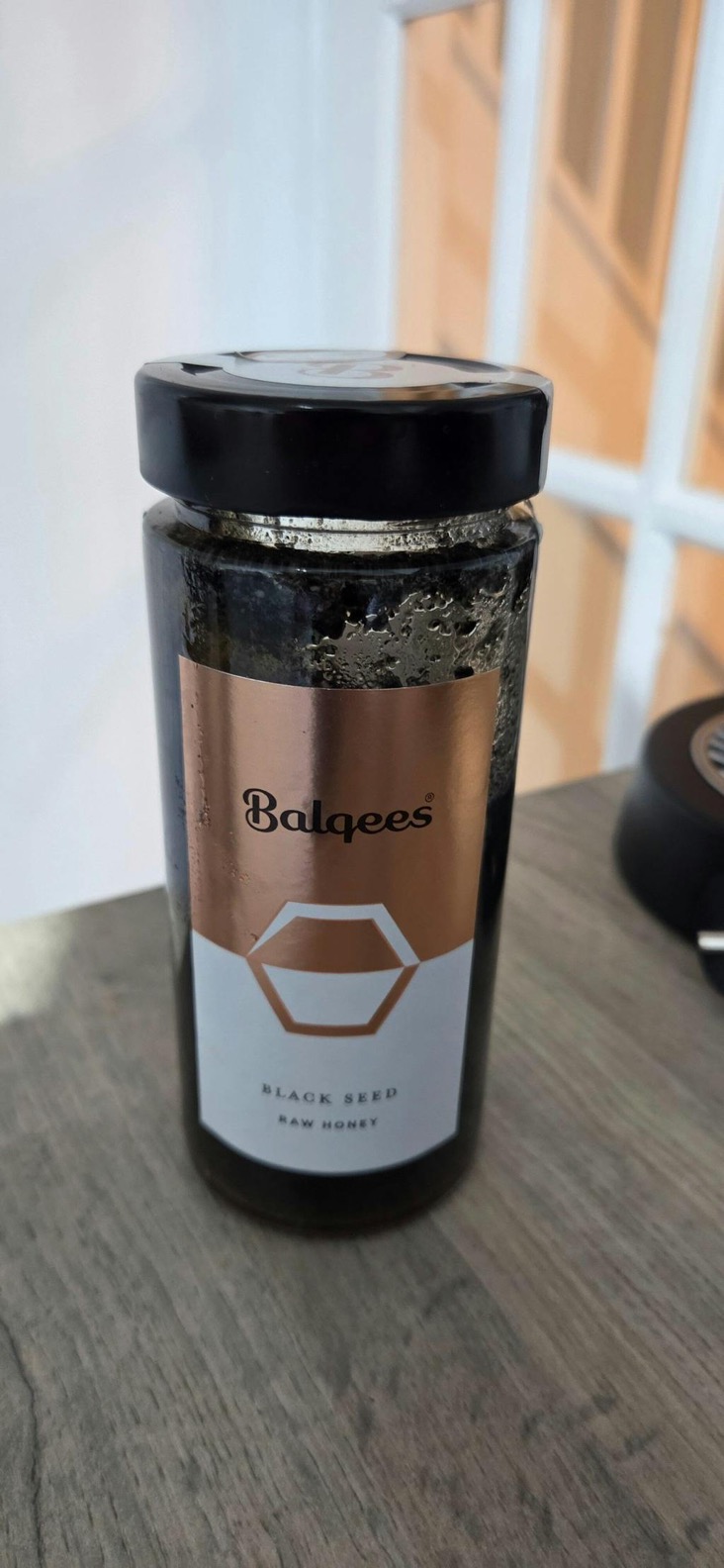 A glass jar of Balqees Black Seed Raw Honey with a black lid, sitting on a wooden surface.