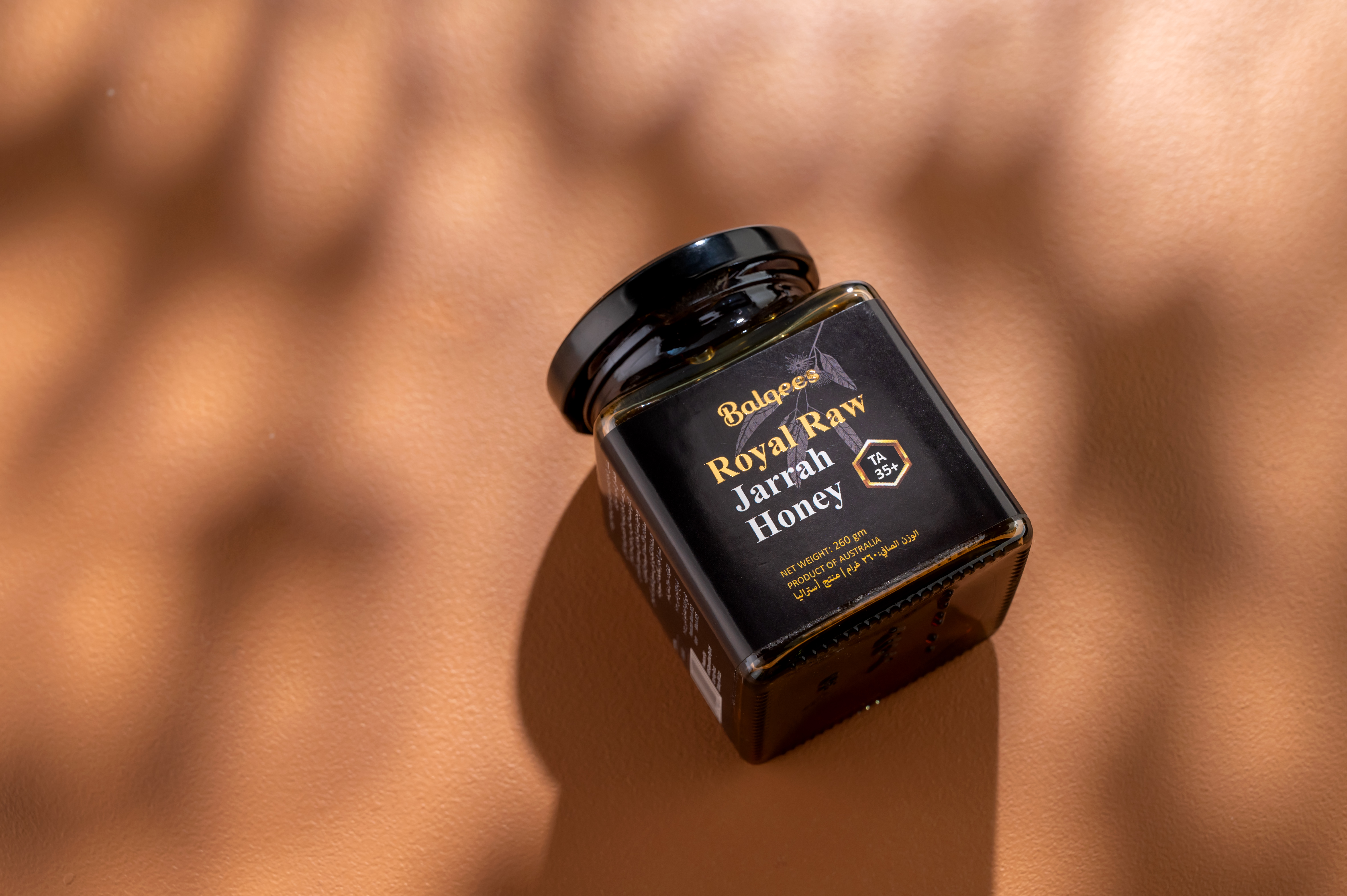 A jar of Balqees Royal Raw Jarrah Honey sits on a textured brown surface with soft shadows.