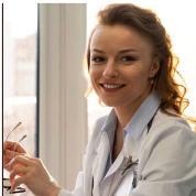 Smiling person in a white coat holding glasses, sitting by a window.