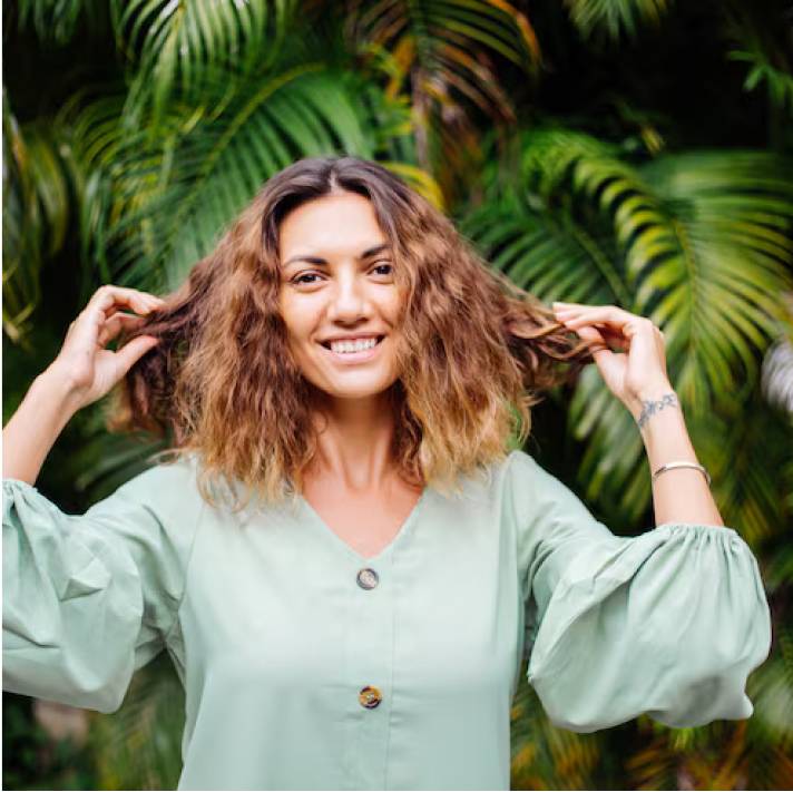 Smiling person with curly hair, wearing a light green blouse, standing in front of palm leaves.