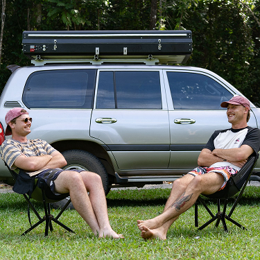 Two men relax in camping chairs in front of a silver SUV with a rooftop tent.