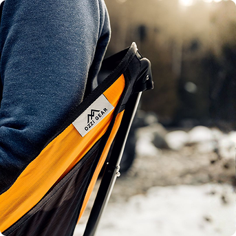 A person sits in an orange and black Ozzi Gear camping chair outdoors during sunset.