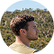 A young man with brown hair in profile, with a background of green, tree-covered hills and blue sky.