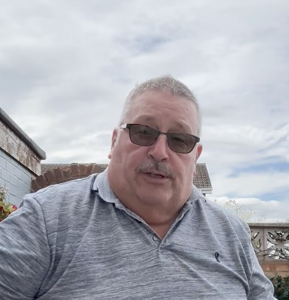 A man with a mustache and sunglasses wearing a gray polo shirt, photographed from a low angle outdoors.