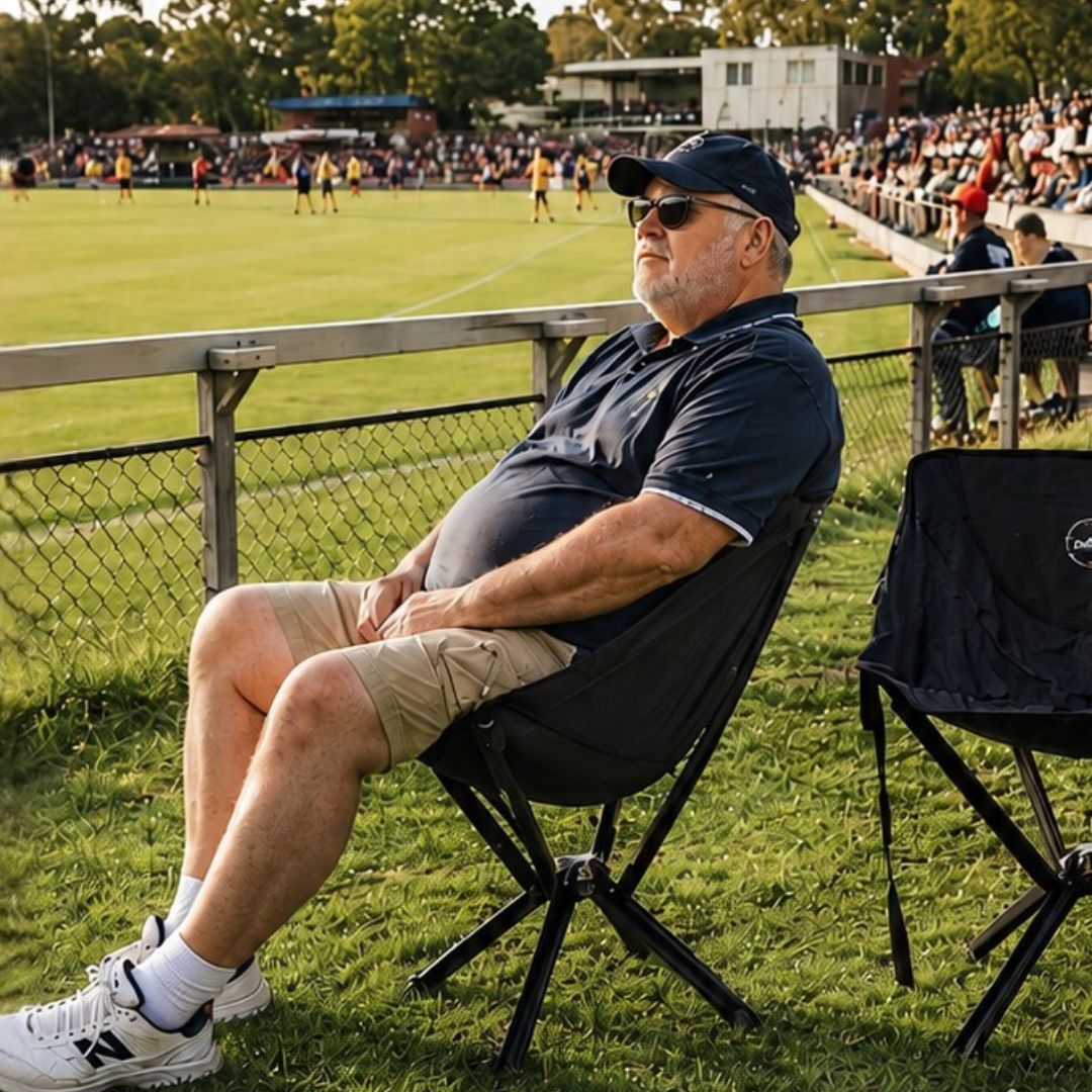 A man in a cap and sunglasses sits in a portable chair on the grass, watching a sporting event.