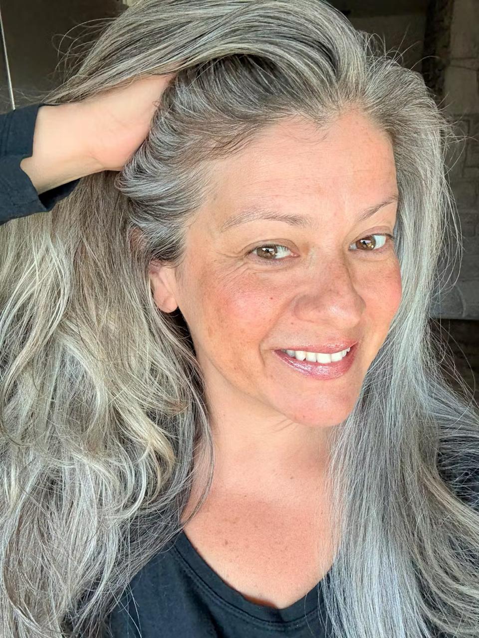 Person smiling, touching their long gray hair, wearing a black top.
