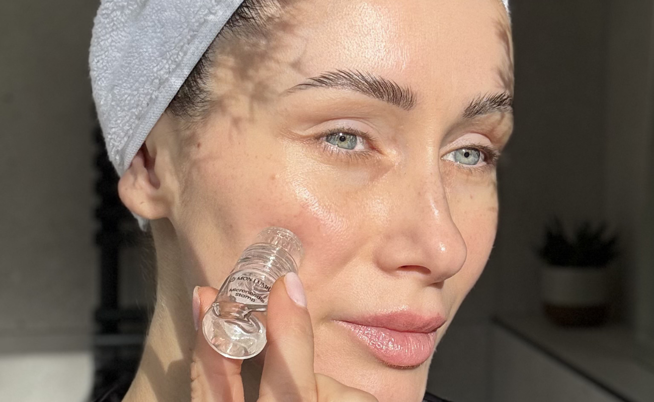A woman with a towel on her head applies a clear liquid from a small vial to her cheek.