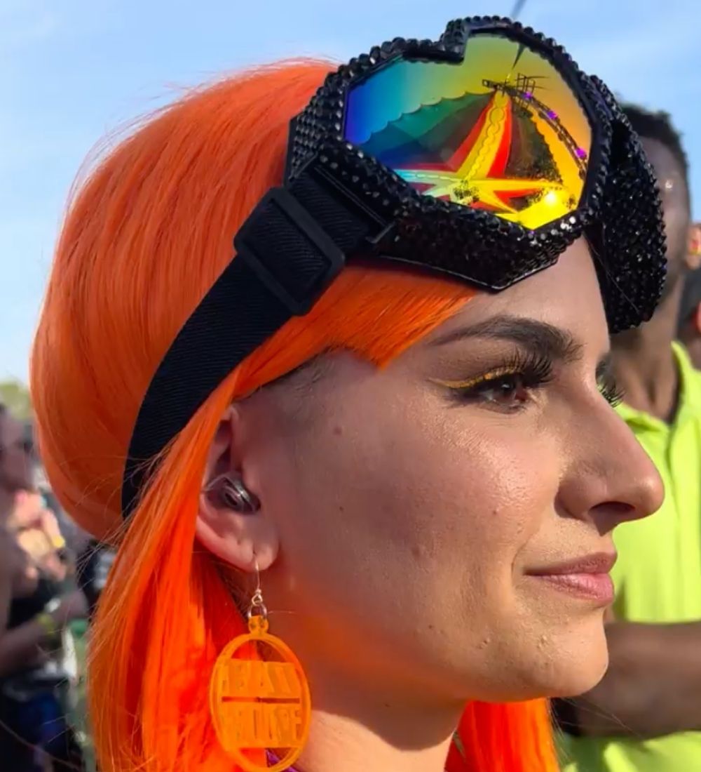 A person with bright orange hair wearing large, bejeweled goggles with iridescent lenses on their forehead.