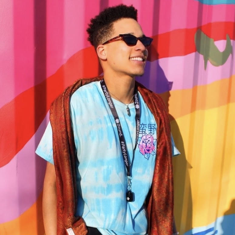 A smiling young man in sunglasses and a tie-dye shirt stands against a colorful striped wall.