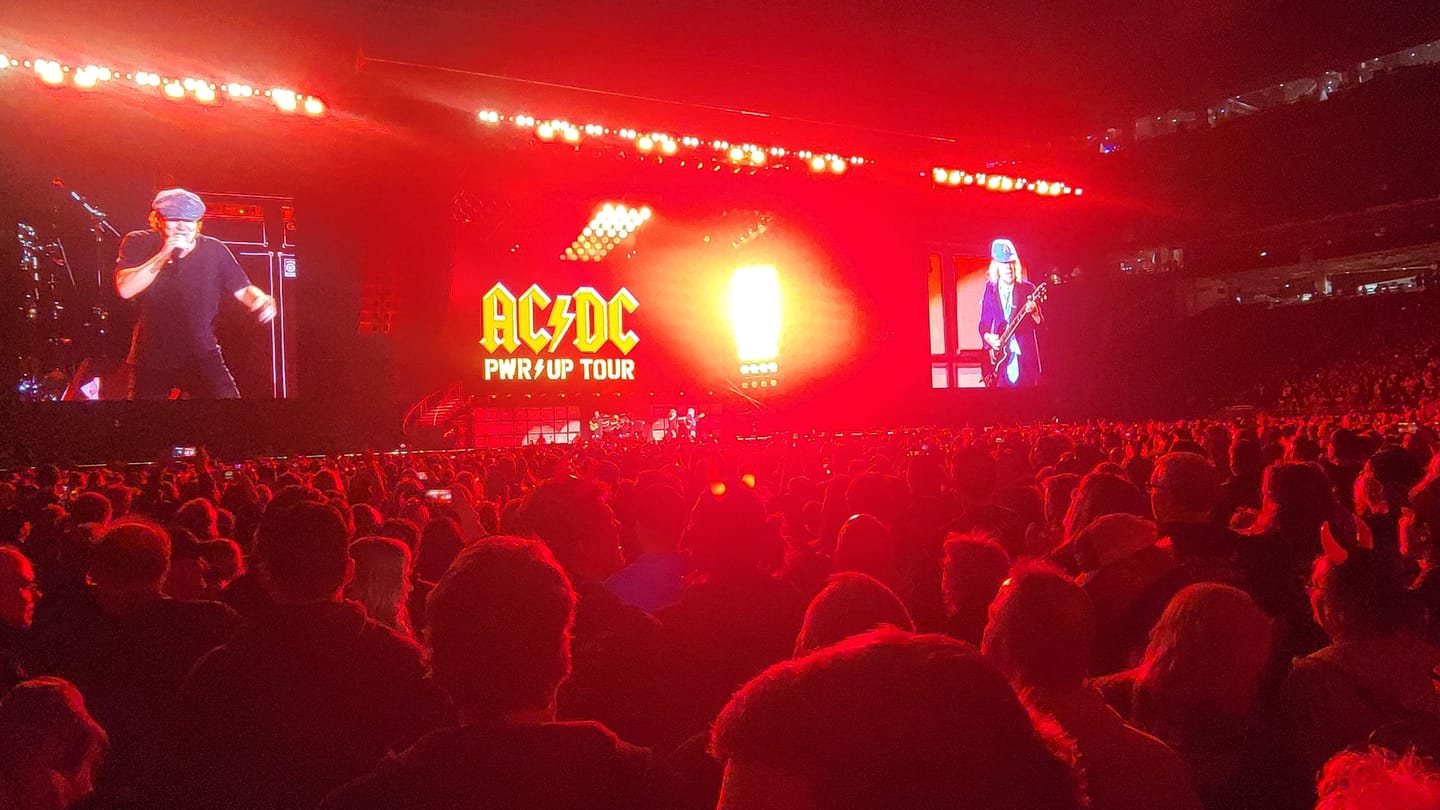 Concert stage with large screens, crowd, and red lighting.