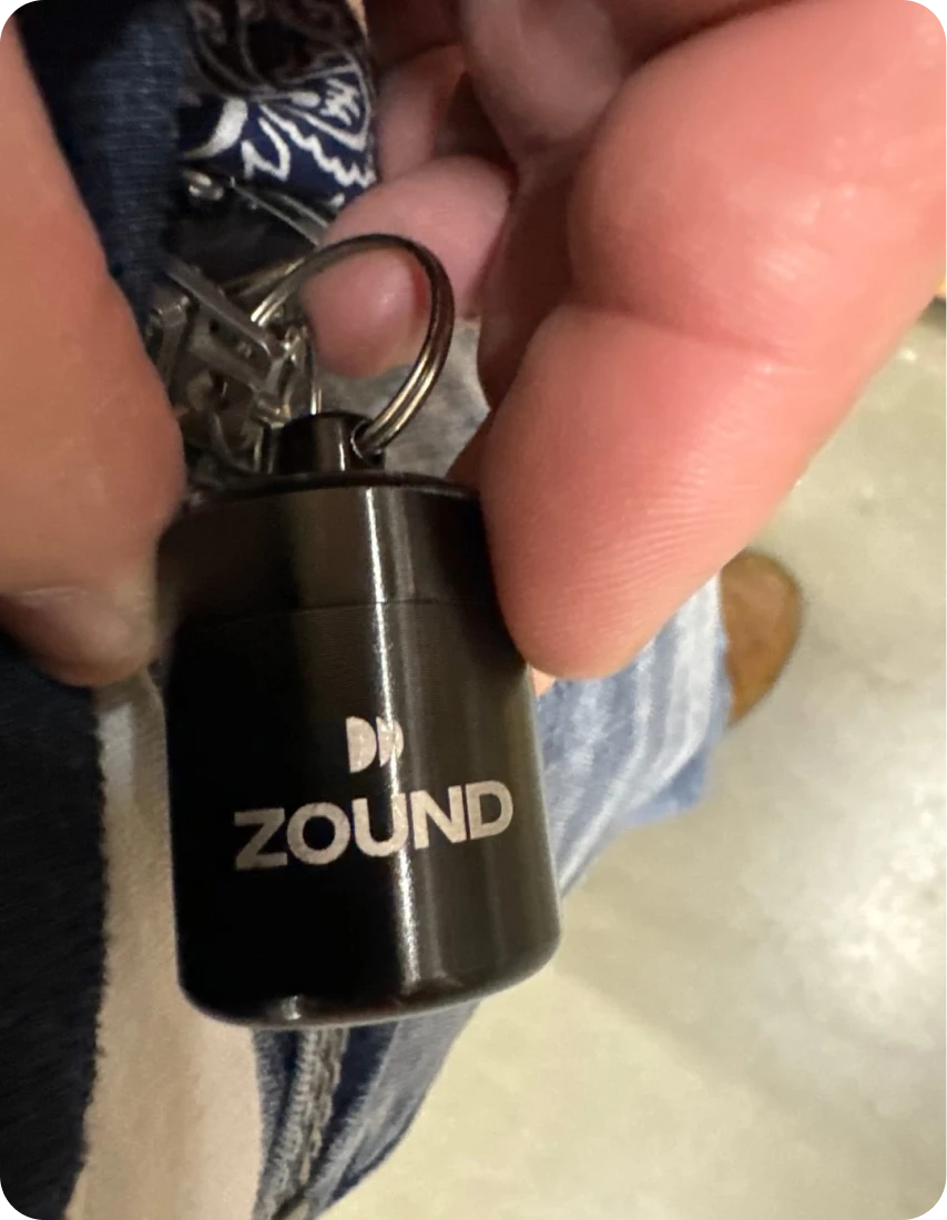 A close-up of a hand holding a small, black cylindrical keychain with the word ZOUND on it.