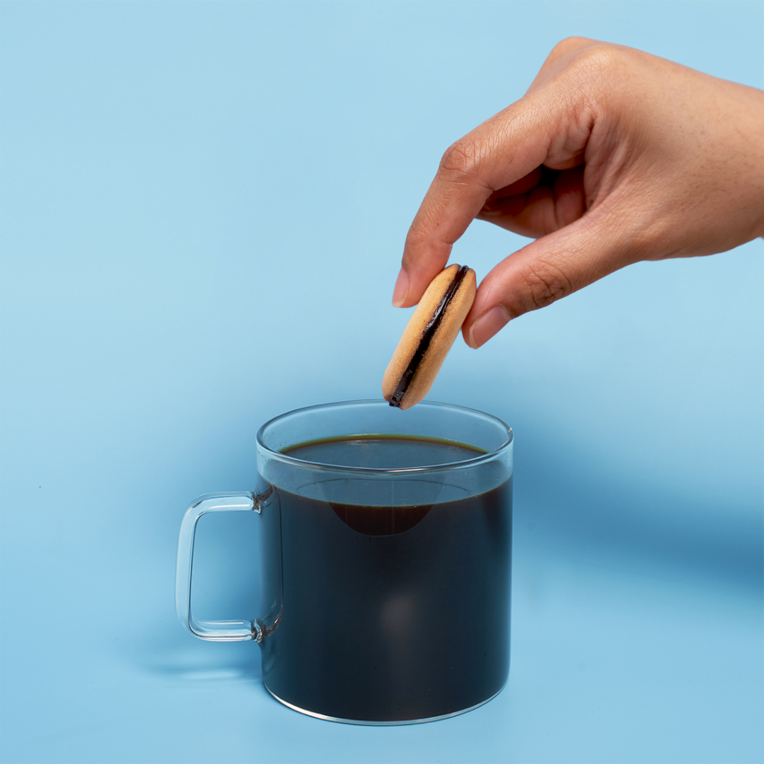 Hand holding a cookie over a glass mug of coffee on a blue background.