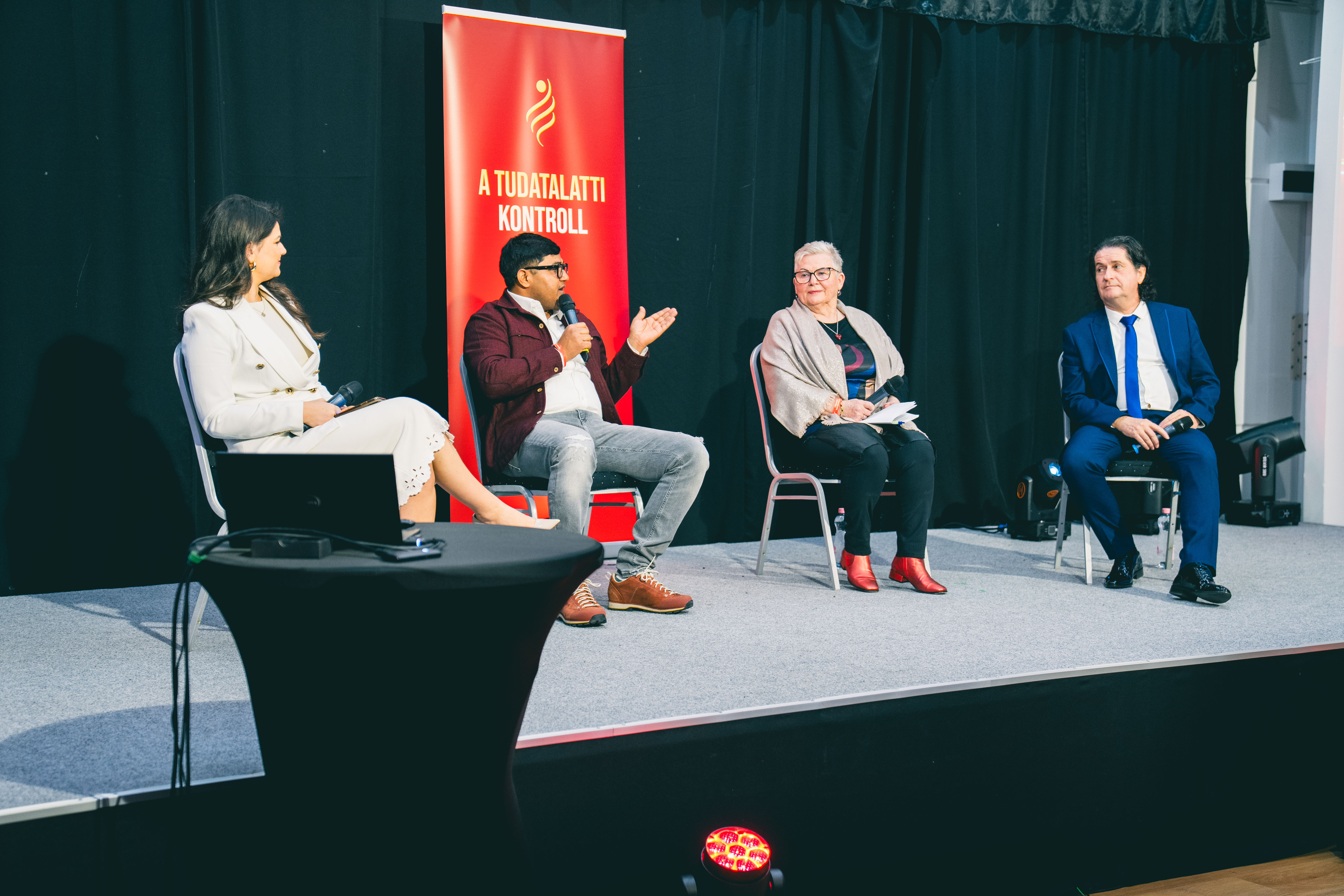 Four people are seated on a stage for a panel discussion; one man is speaking into a microphone.