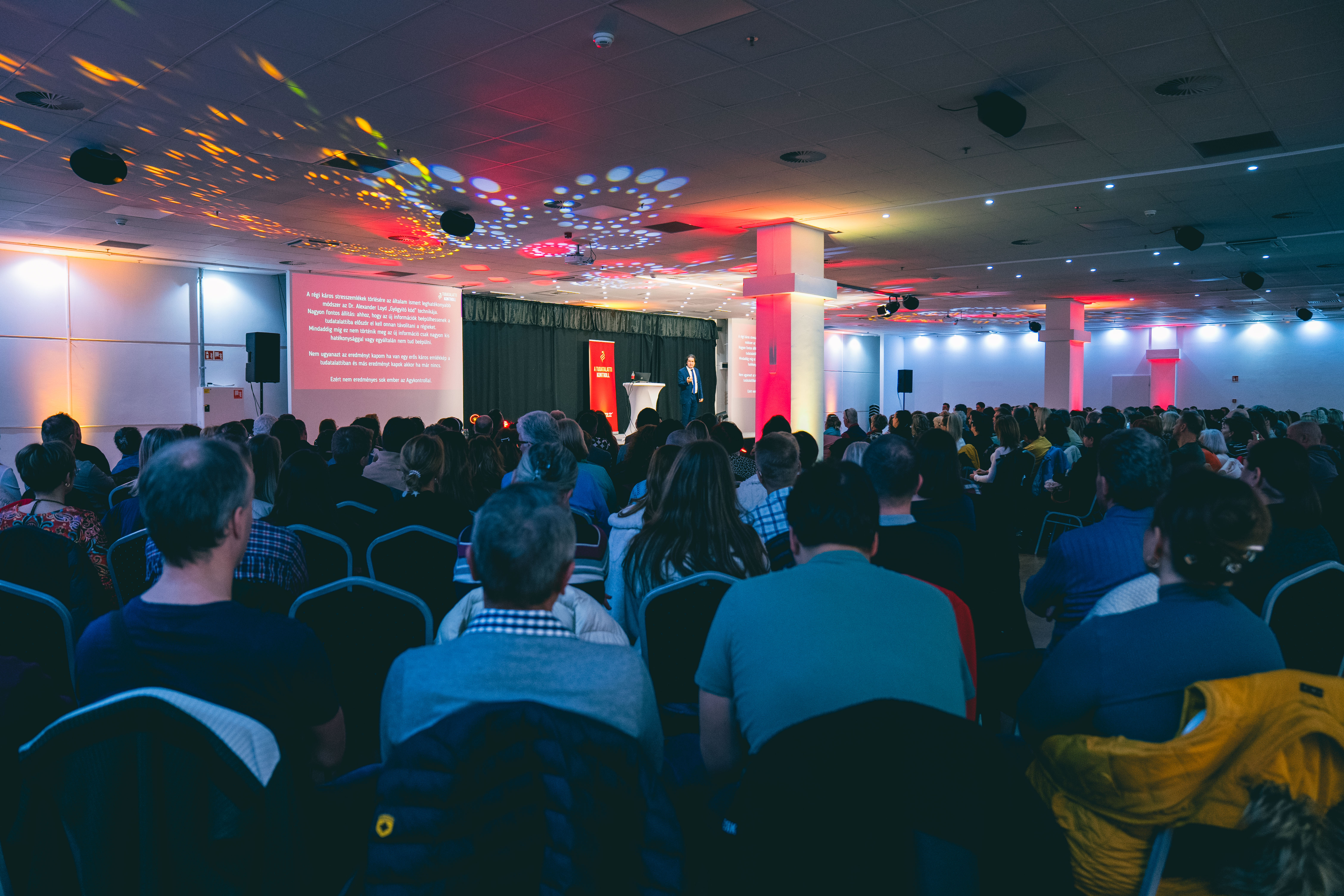A large audience watches a speaker on stage giving a presentation in a conference hall.