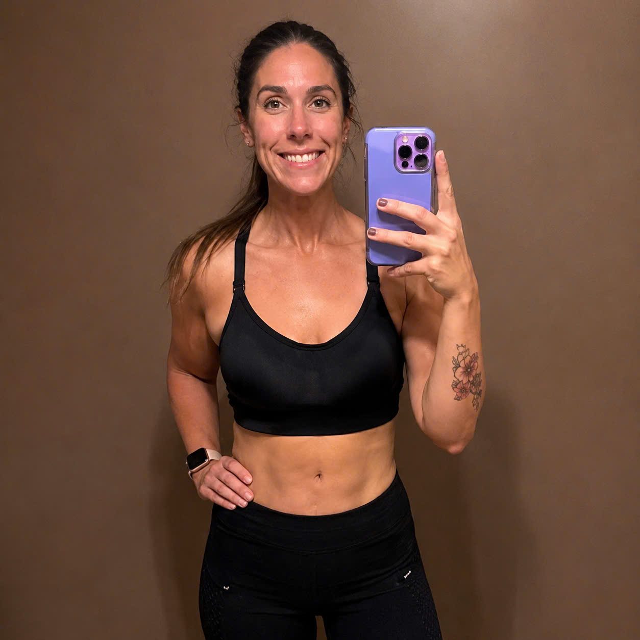 A smiling woman in a black sports bra and leggings takes a mirror selfie with her phone.