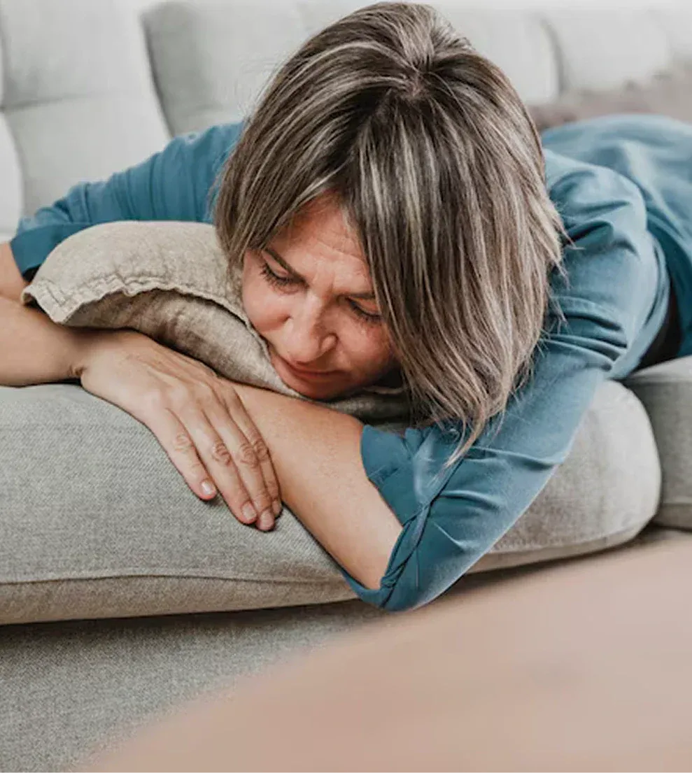 A woman in a blue shirt lies face down on a gray couch with her eyes closed.