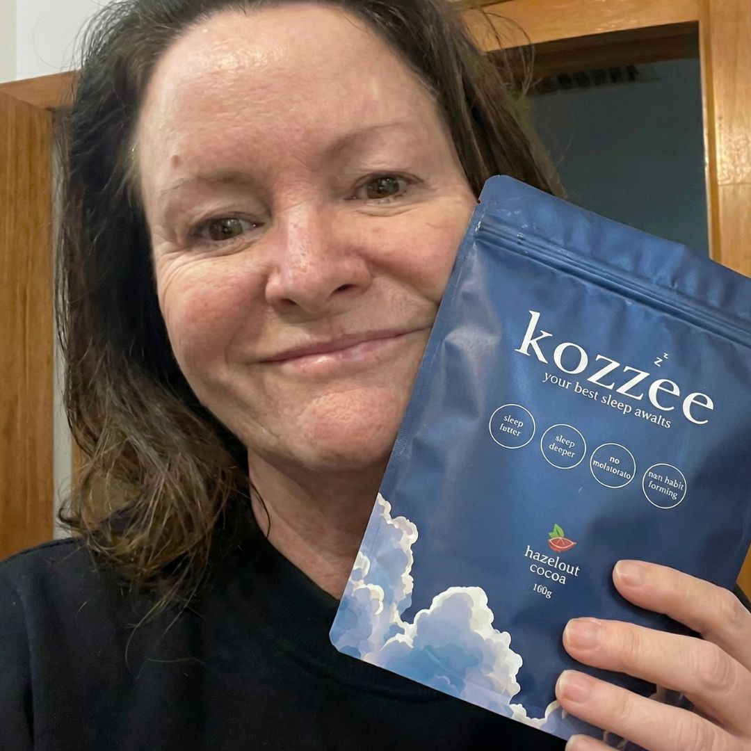 A smiling woman holds up a blue pouch of Kozzee hazelnut cocoa sleep aid.
