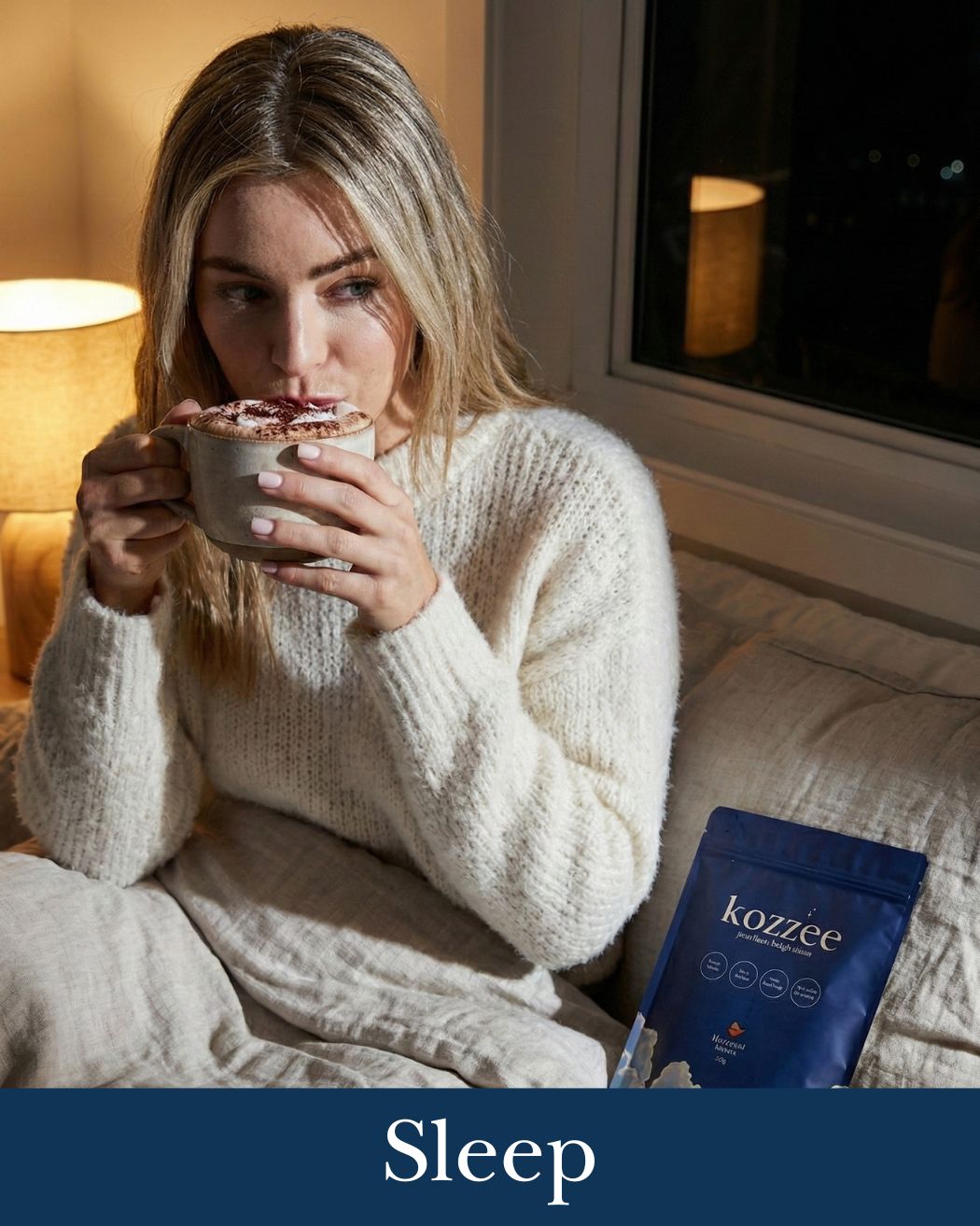 A woman in a cozy white sweater sips a drink from a mug while sitting in bed at night.