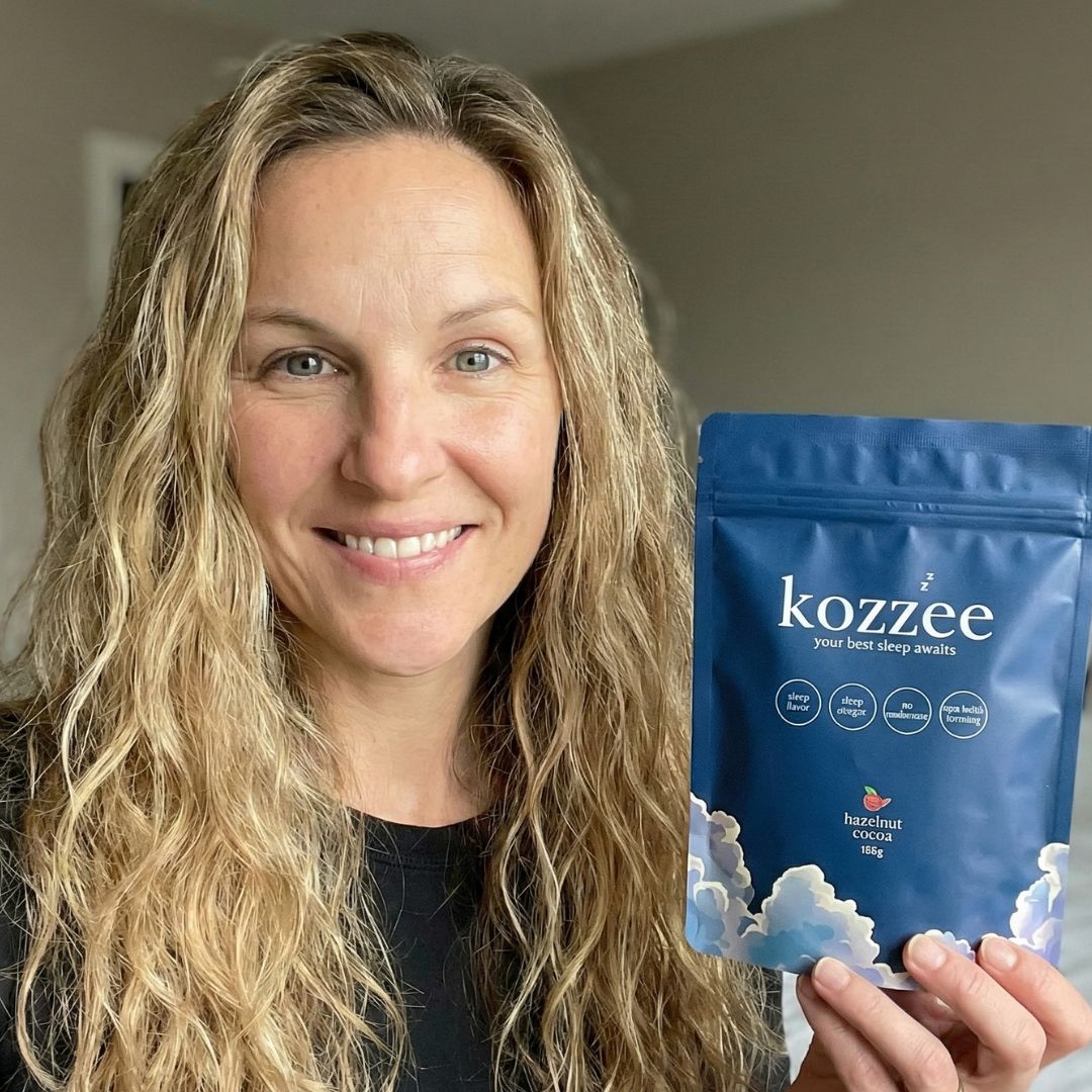 A smiling woman with long blonde hair holds up a blue pouch of Kozzee hazelnut cocoa.